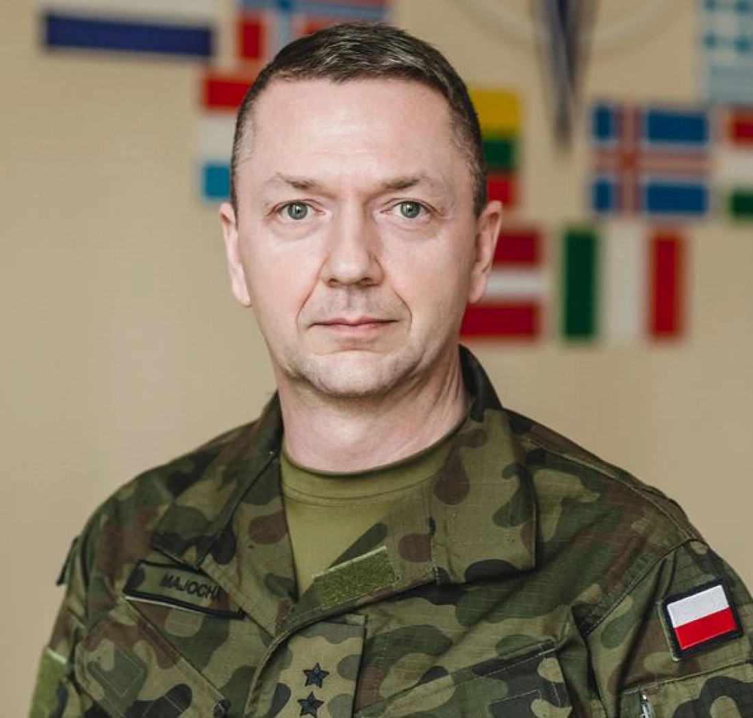 Mężczyzna w mundurze wojskowym, stojący przed tłem z flagami.