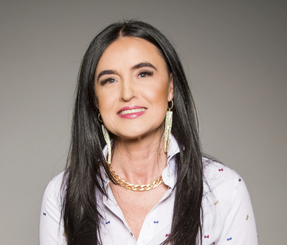 Kobieta o długich, ciemnych włosach, uśmiechająca się, ubrana w białą koszulę ozdobioną kolorowymi detalami i złote biżuterie.