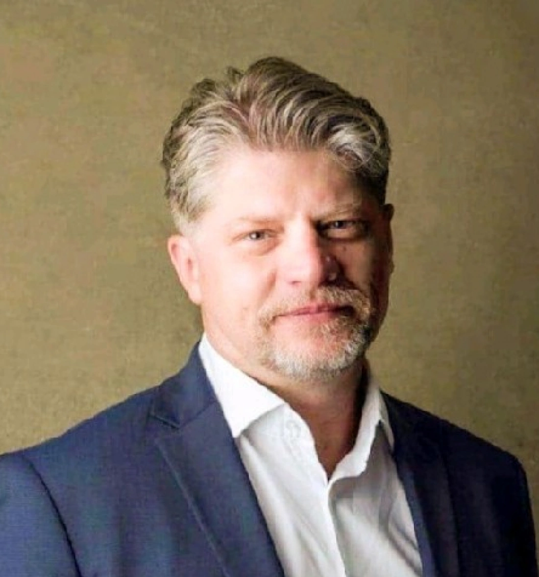 Mężczyzna w garniturze, z uśmiechem na twarzy, stojący w neutralnym tle.