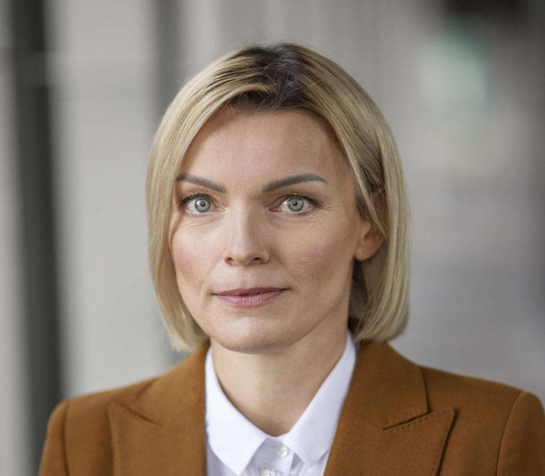 Portret kobiety w eleganckim ubraniu, z krótkimi blond włosami i poważnym wyrazem twarzy.