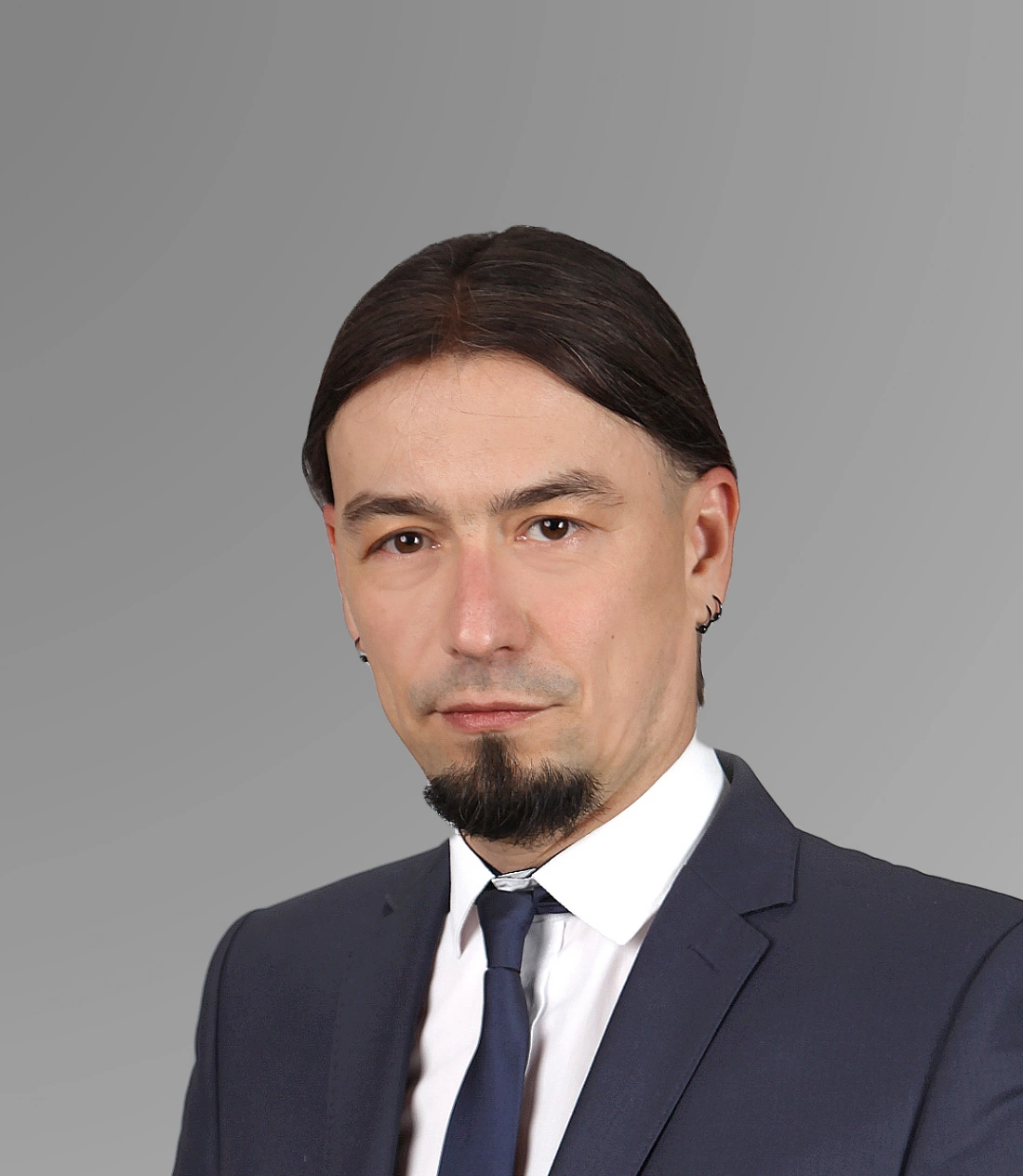 Mężczyzna w garniturze z ciemnymi włosami i brodą, stojący na szarym tle.