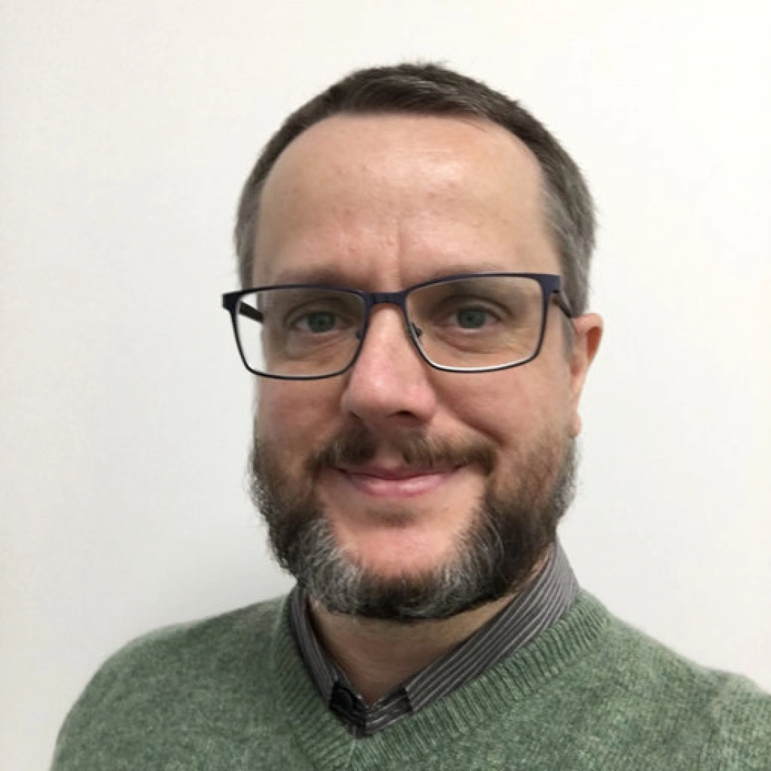 Mężczyzna z brodą i okularami, uśmiechający się na tle jasnej ściany.