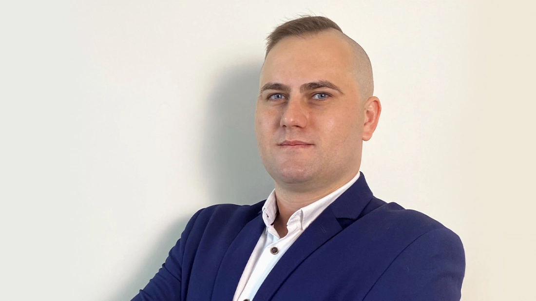 Mężczyzna w garniturze, stojący na neutralnym tle i patrzący pewnie w obiektyw.