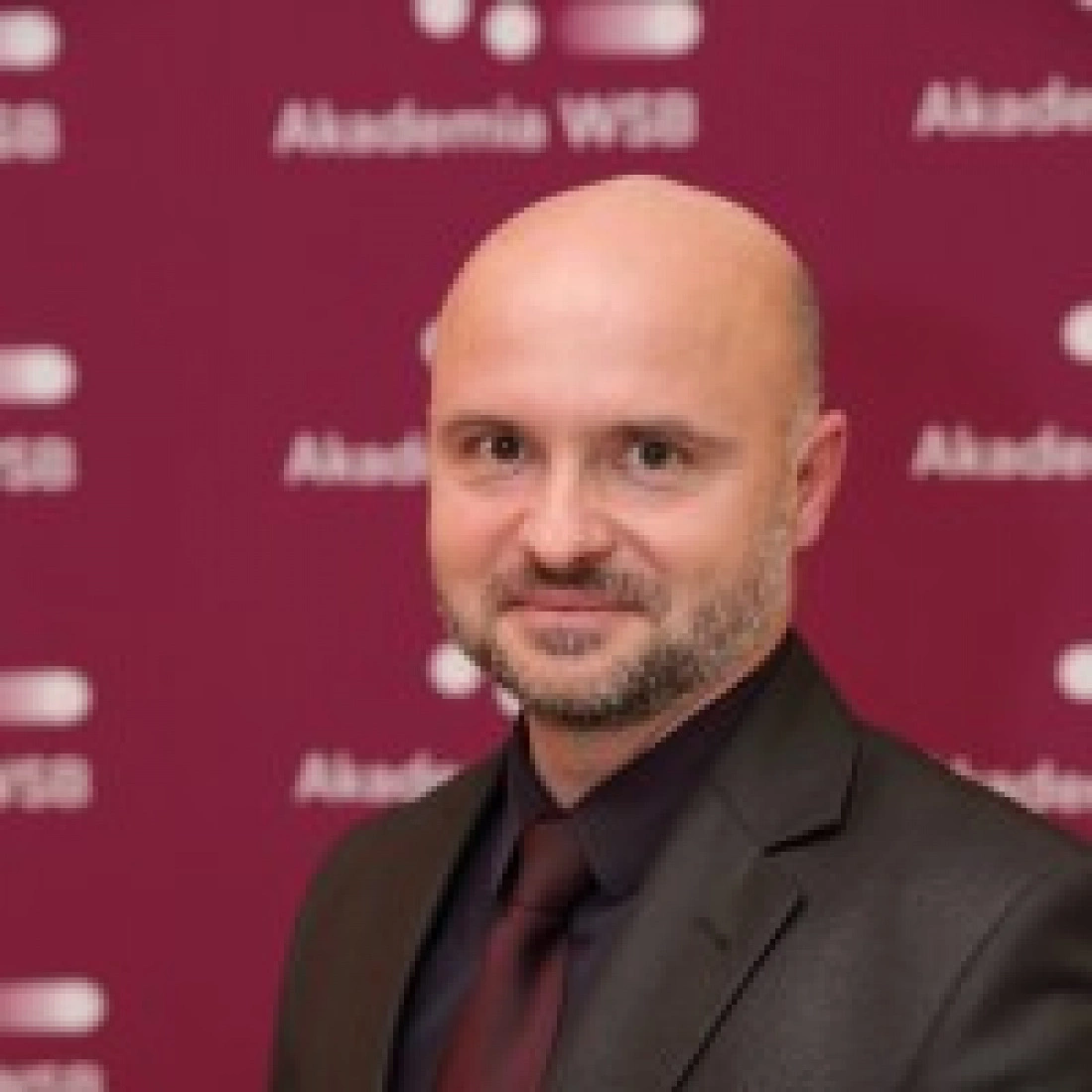 Mężczyzna w eleganckim garniturze, pozujący przed tłem z logo Akademii WSB.