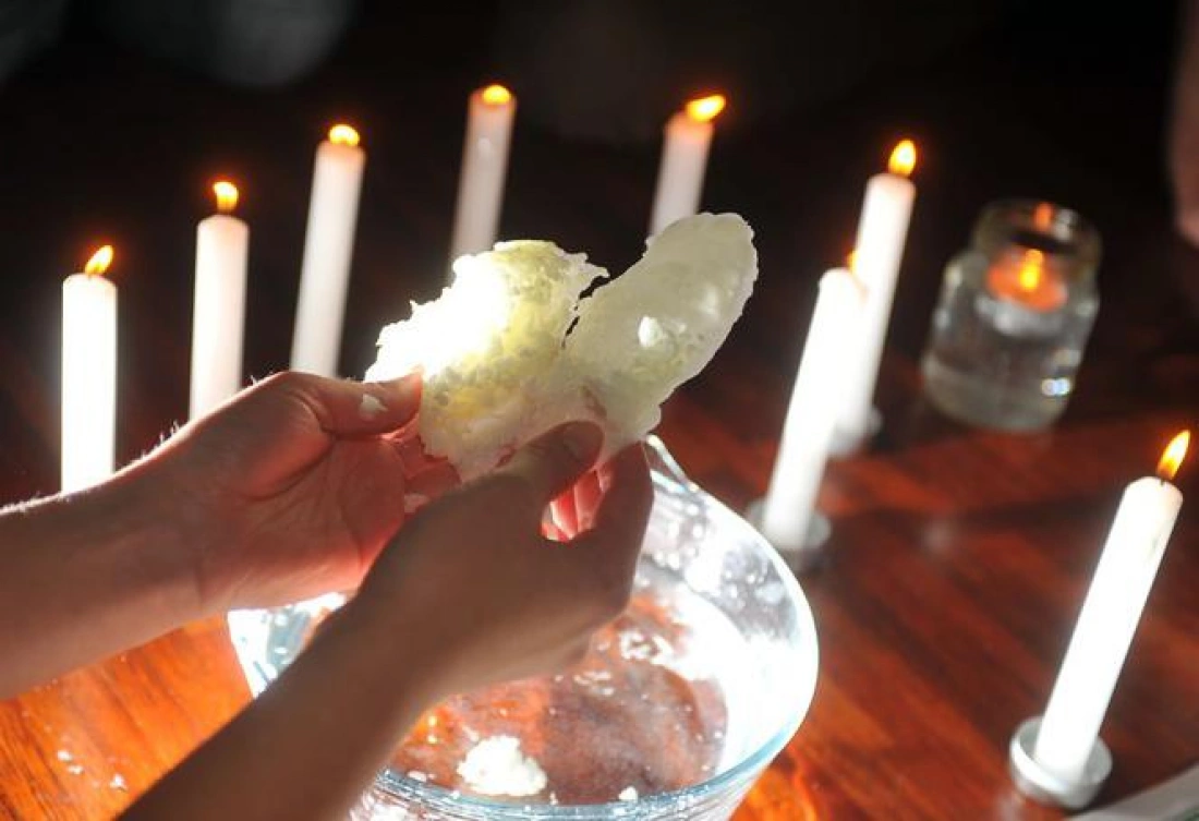 Osoba trzyma w dłoniach formę woskową nad miską wypełnioną wodą, otoczoną zapalonymi świecami.