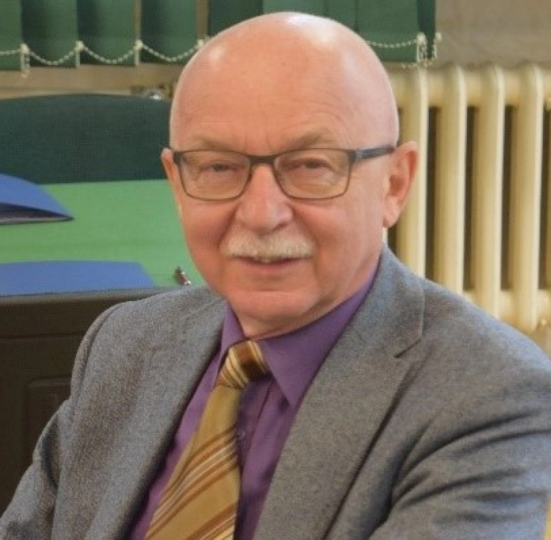 Mężczyzna w okularach, uśmiechający się, w eleganckim garniturze, siedzi przy stole.