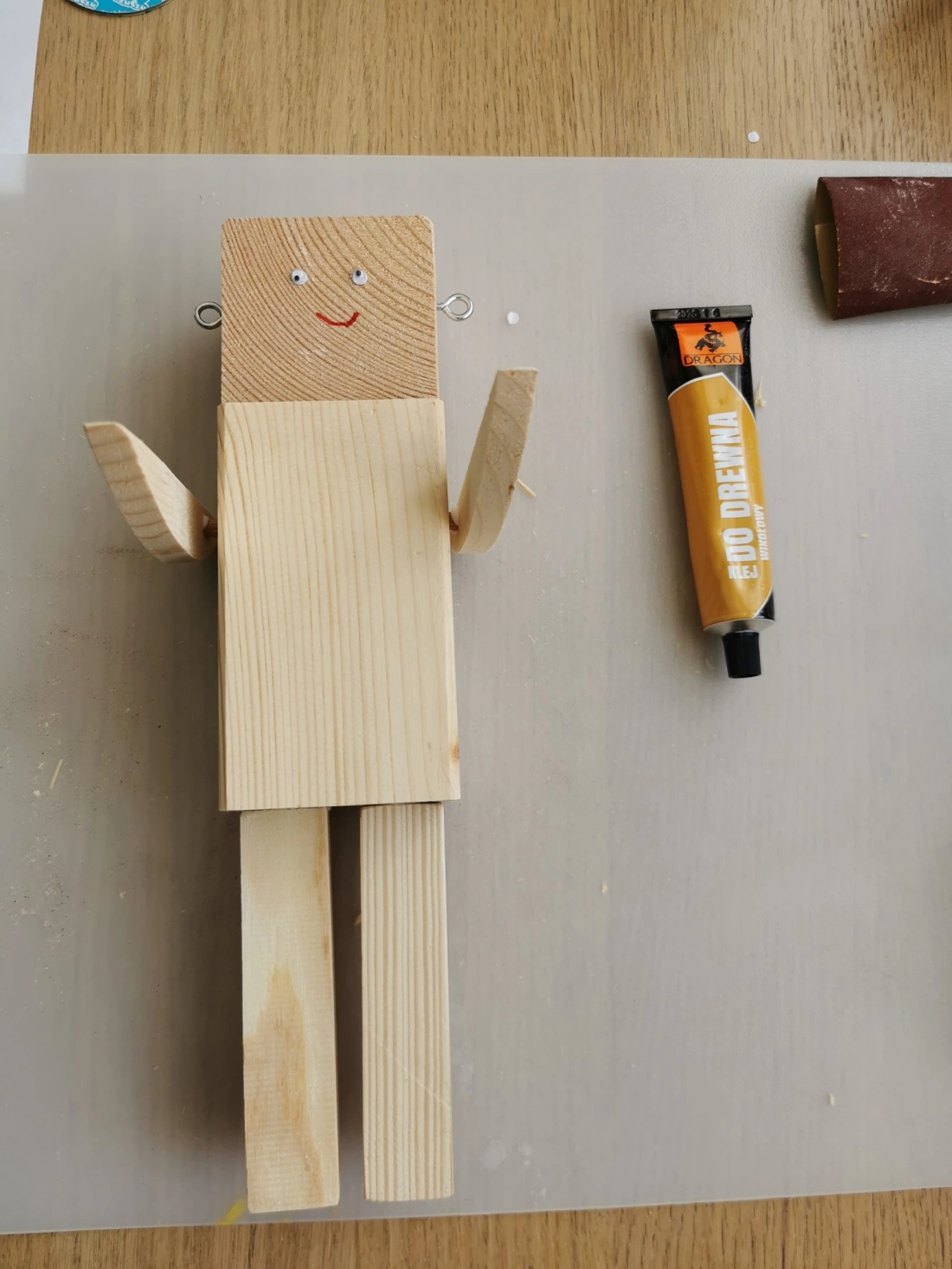 Drewniana postać z uśmiechem, z ruchomymi ramionami, obok tubka kleju i papier ścierny.