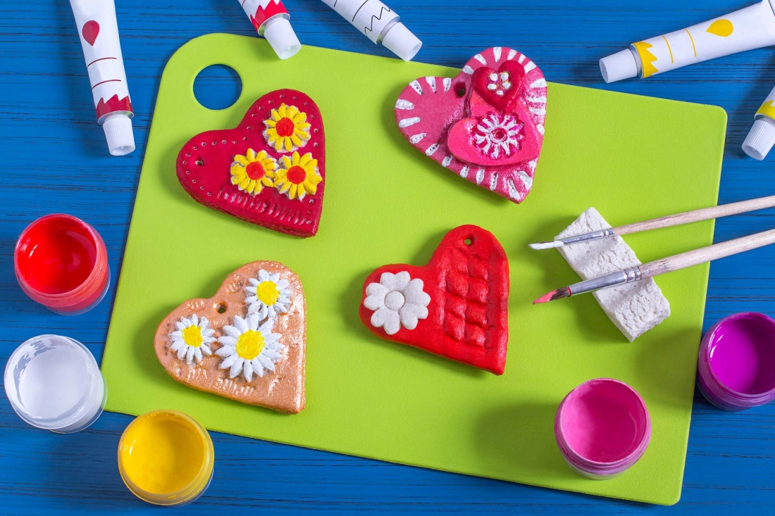 Kreatywne serca w różnych kolorach i wzorach na zielonej desce, otoczone farbami i pędzlami.