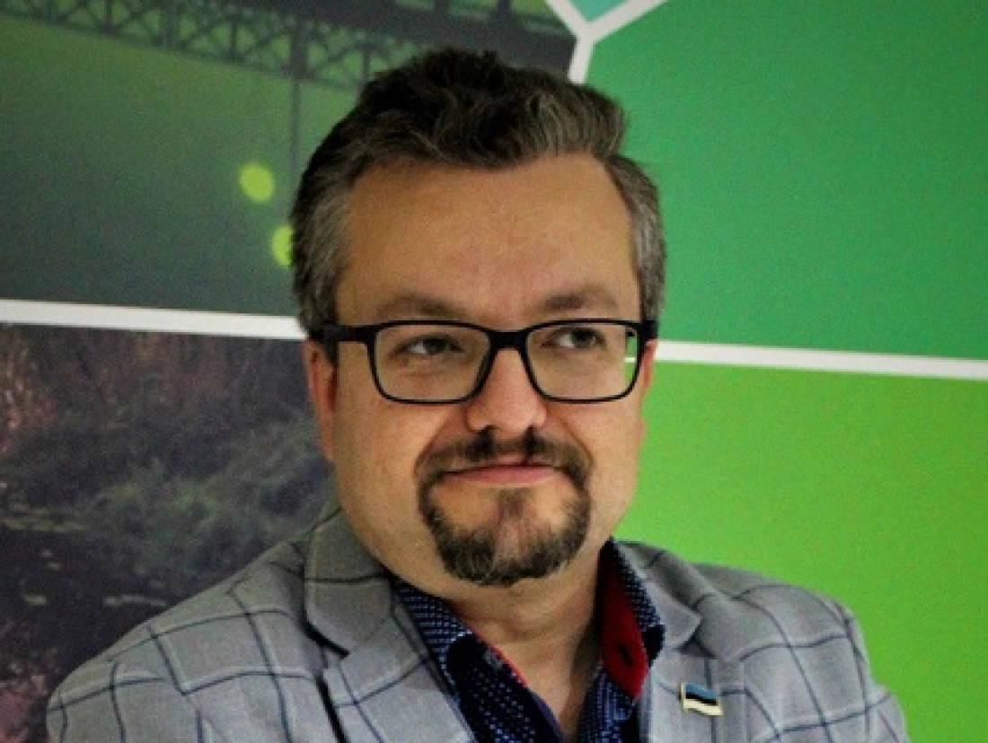 Mężczyzna w okularach, uśmiechający się, w szarym garniturze na tle zielonej ściany.