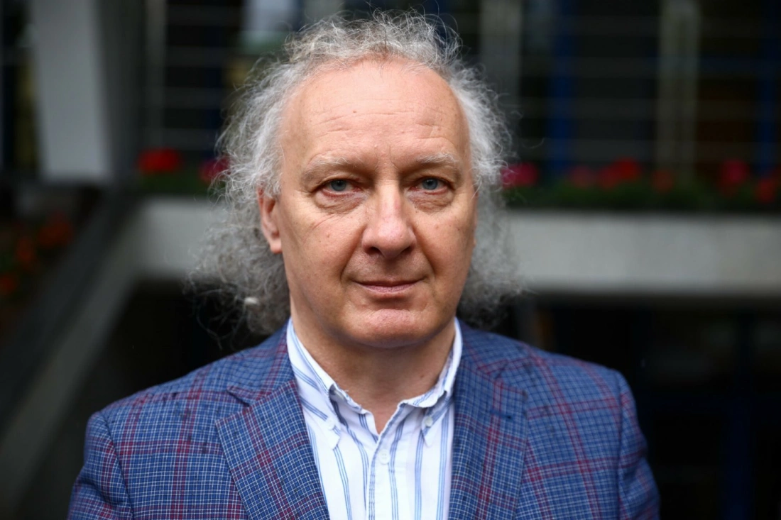 Mężczyzna z długimi, kręconymi włosami, ubrany w niebieską marynarkę w kratę, stoi na tle budynku.