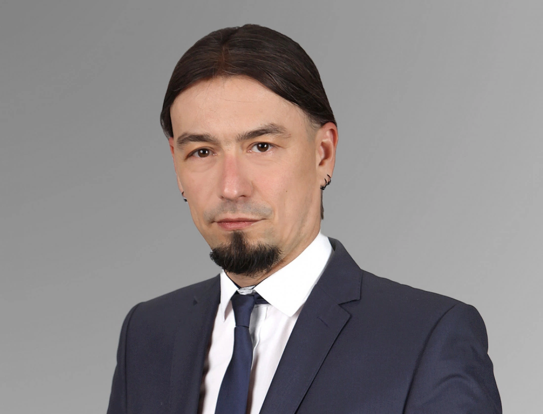 Mężczyzna w eleganckim garniturze, z kr&oacute;tkimi włosami i zarostem, na neutralnym tle.