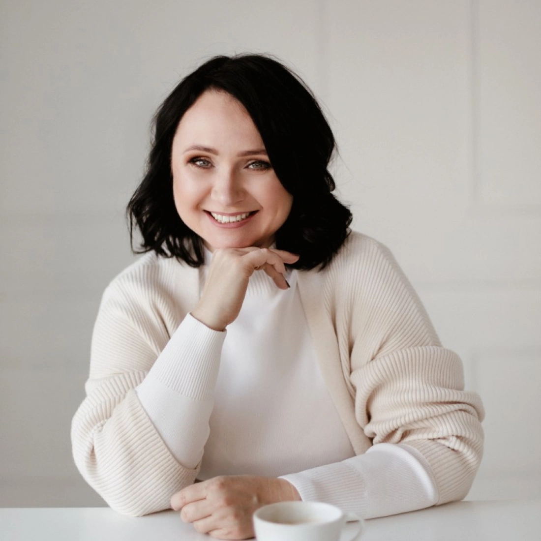 Uśmiechnięta kobieta siedzi przy stole, opierając rękę na brodzie, z filiżanką kawy przed sobą.