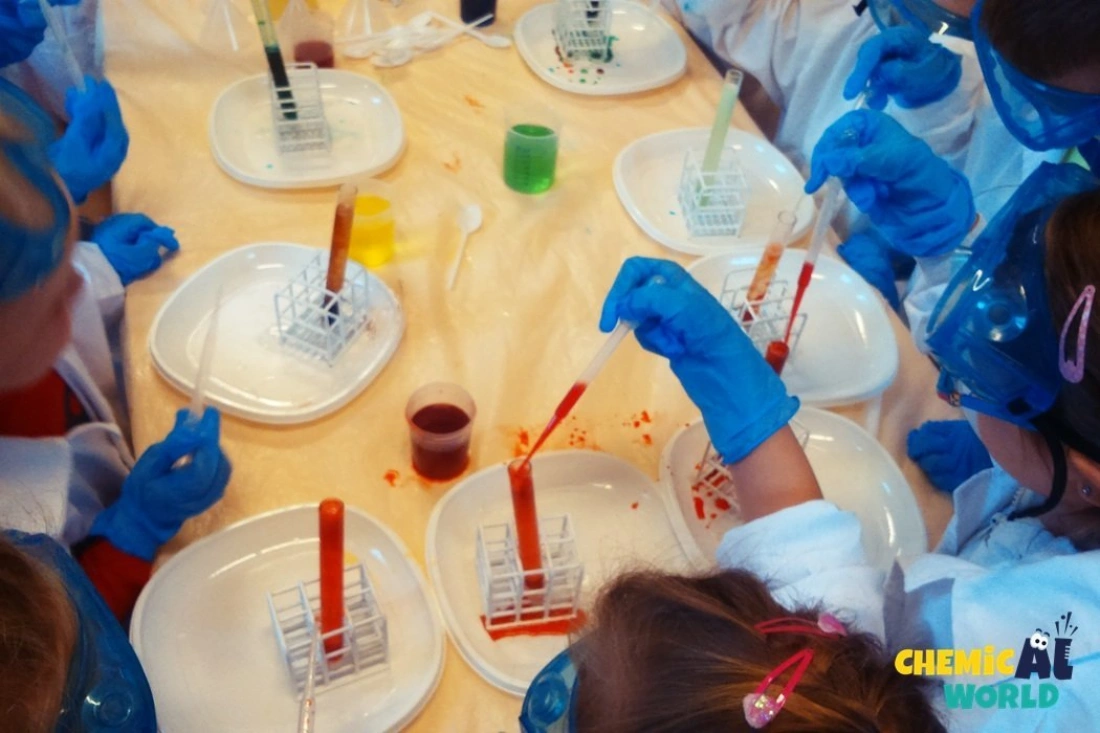 Grupa dzieci w kombinezonach laboratoryjnych wykonuje eksperymenty chemiczne, używając probówek i pipet.