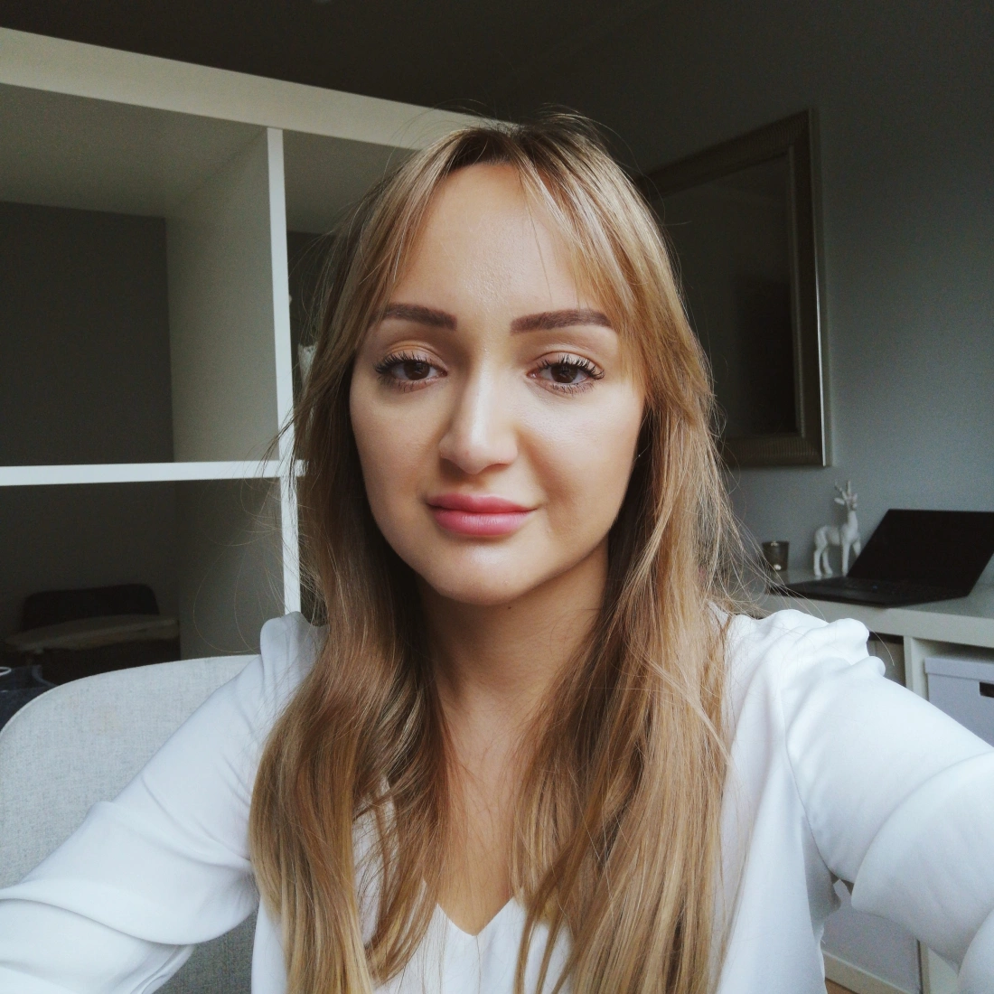 Kobieta z długimi, jasnymi włosami, w białej bluzce, uśmiecha się do kamery w jasnym pomieszczeniu.