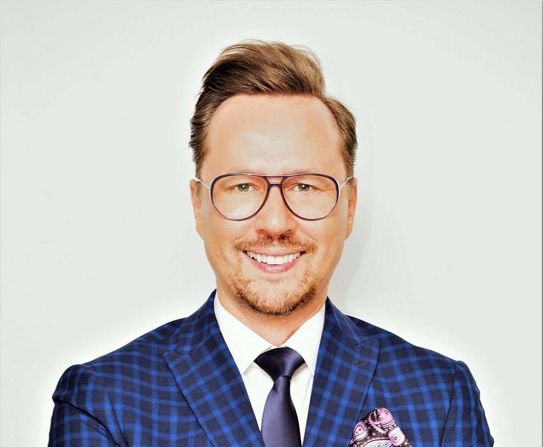 Mężczyzna w eleganckim garniturze z okularami, uśmiechający się na jednolitym tle.