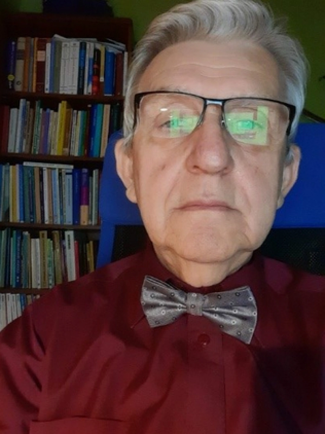 Mężczyzna w okularach i eleganckiej koszuli z muszką, siedzący przed regałem pełnym książek.