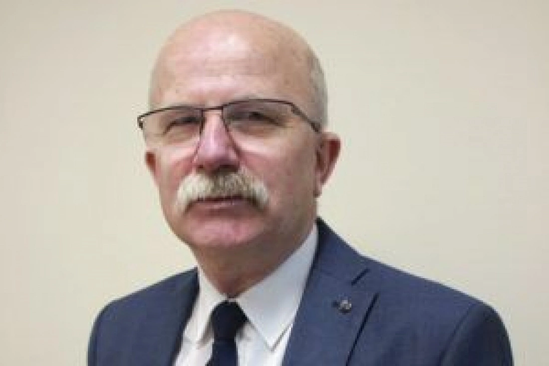 Mężczyzna w okularach z siwą bródką, ubrany w garnitur, na jasnym tle.