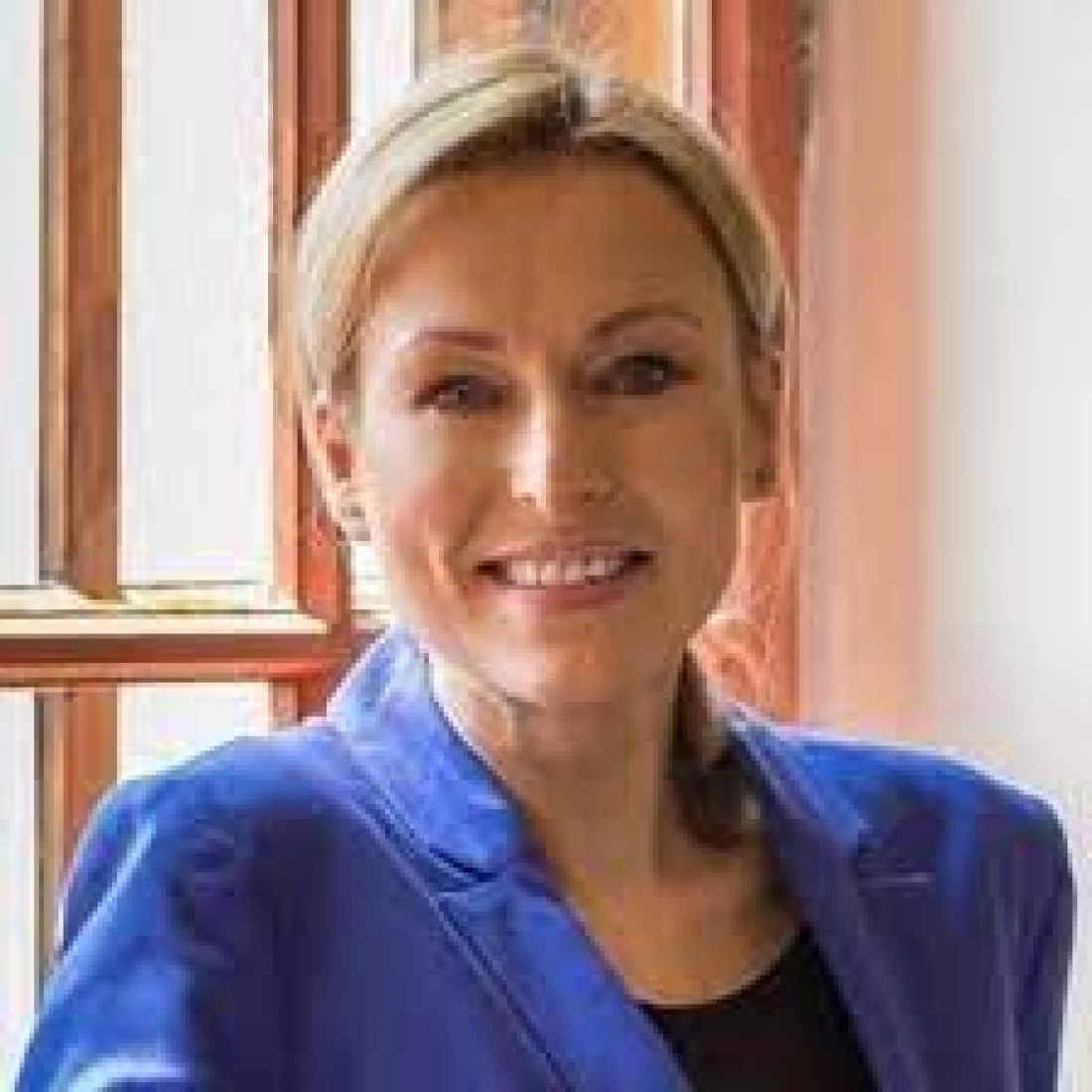 Kobieta w niebieskiej marynarce, uśmiechająca się przy oknie.