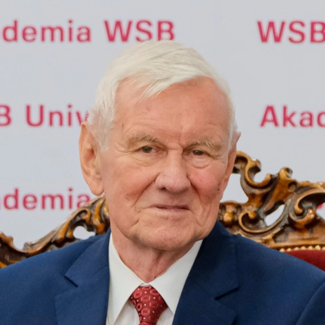 Starszy mężczyzna w garniturze, siedzący na eleganckim krześle, z uśmiechem na tle banneru Akademii WSB.