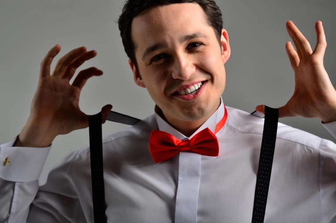 Mężczyzna w białej koszuli i czerwoną muszką uśmiecha się, trzymając szelki.