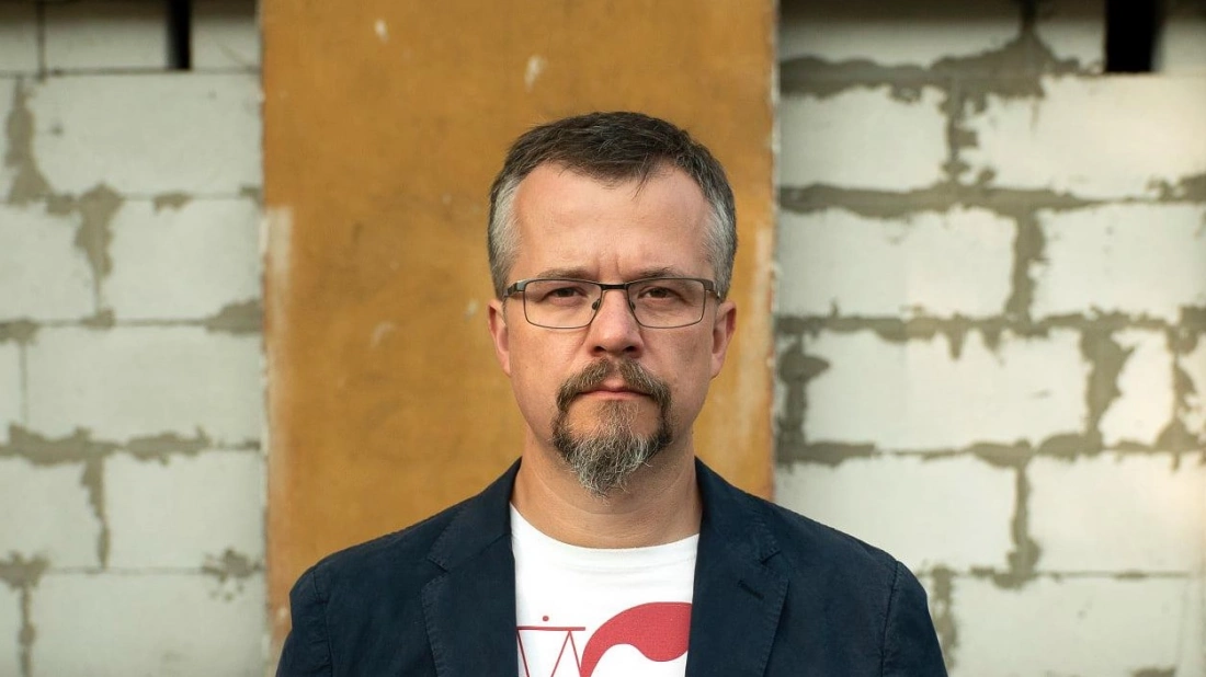 Mężczyzna w okularach i z brodą, ubrany w marynarkę, stoi przed nier&oacute;wną ścianą.