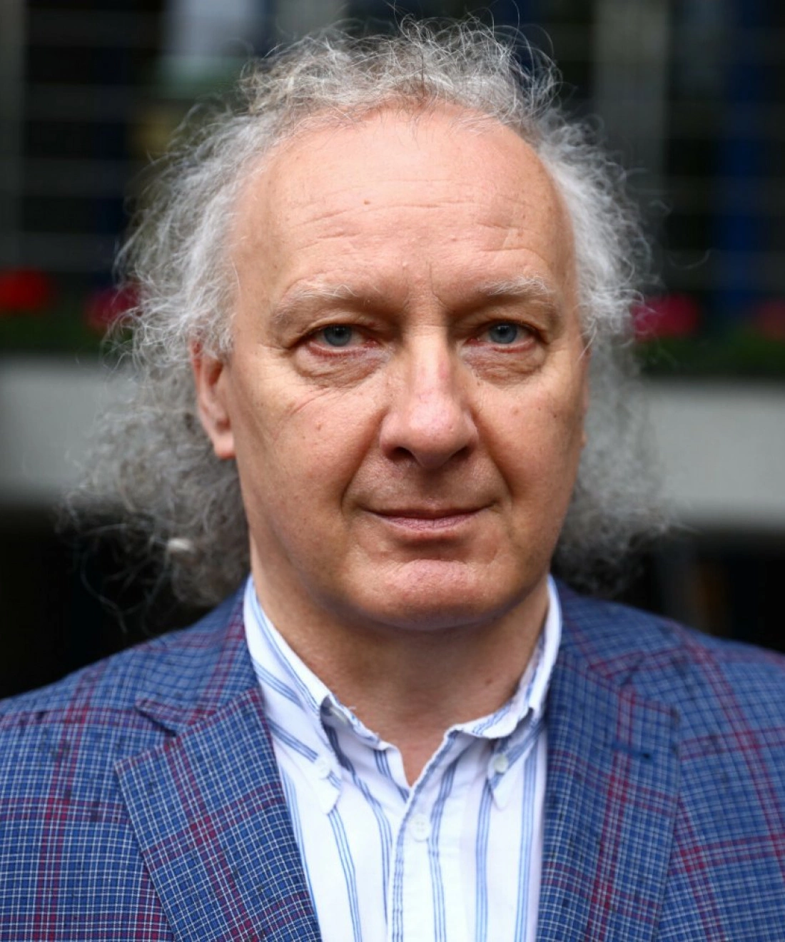 Mężczyzna z kręconymi włosami i niebieskimi oczami, ubrany w niebieską marynarkę w kratę i białą koszulę w paski.