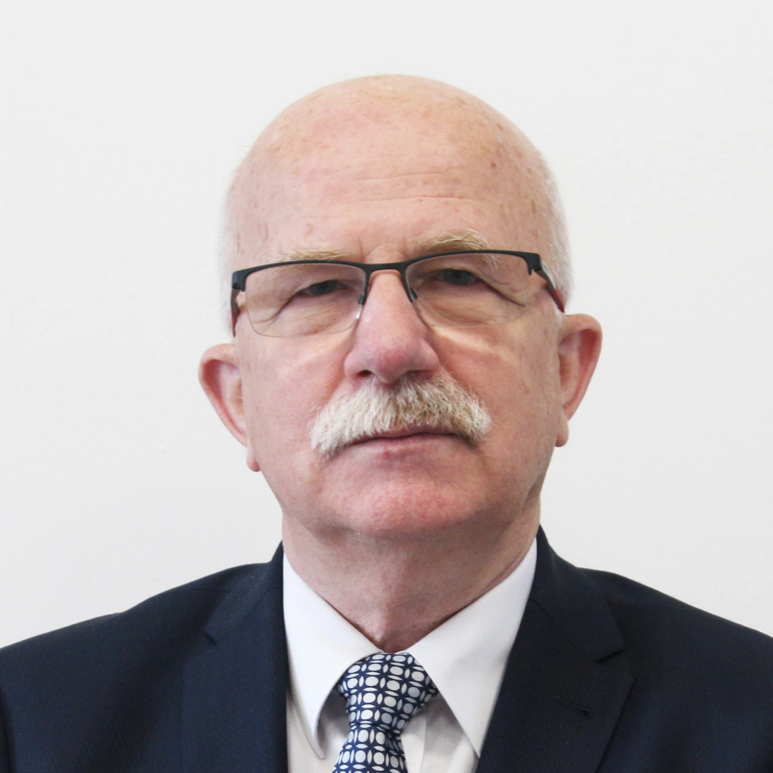 Mężczyzna z okularami i siwymi włosami, w garniturze, na tle białej ściany.