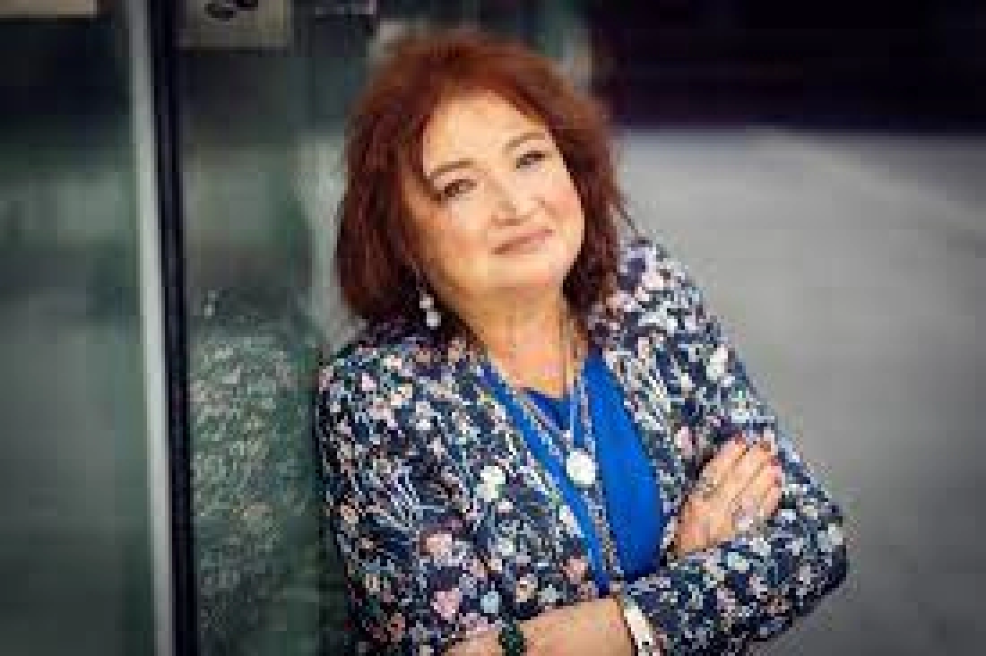 Kobieta o rudych włosach uśmiecha się, stojąc w eleganckiej bluzce i kwiecistej marynarce.