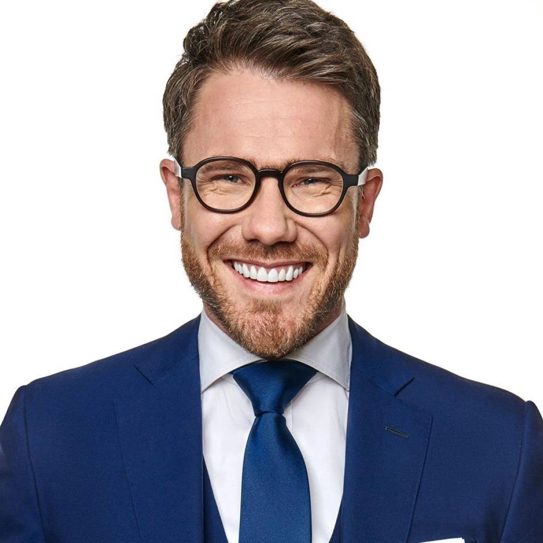 Mężczyzna w okularach, ubrany w niebieski garnitur, uśmiecha się szeroko.