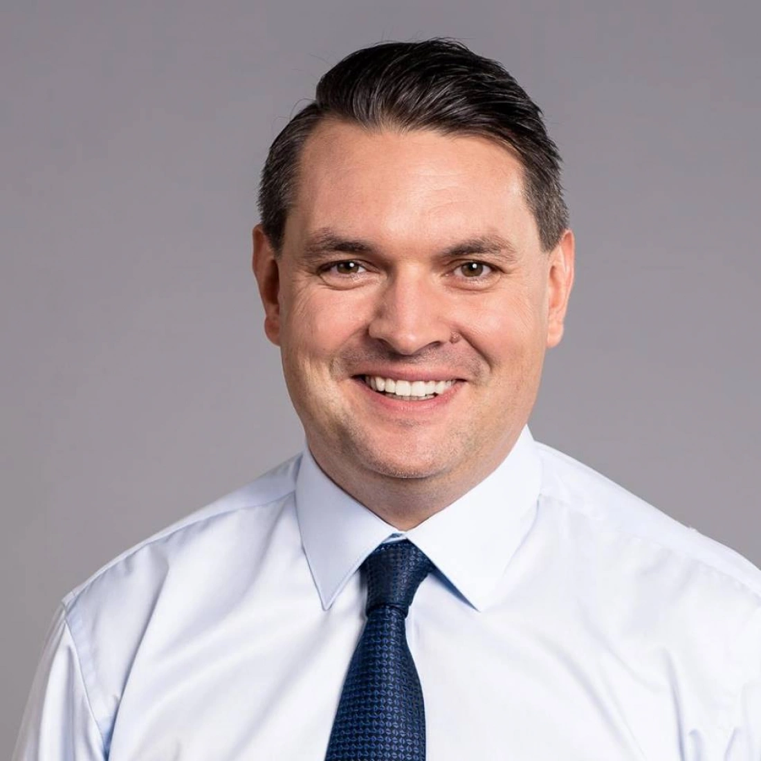 Mężczyzna w białej koszuli i granatowym krawacie, uśmiechający się na tle szarej ściany.