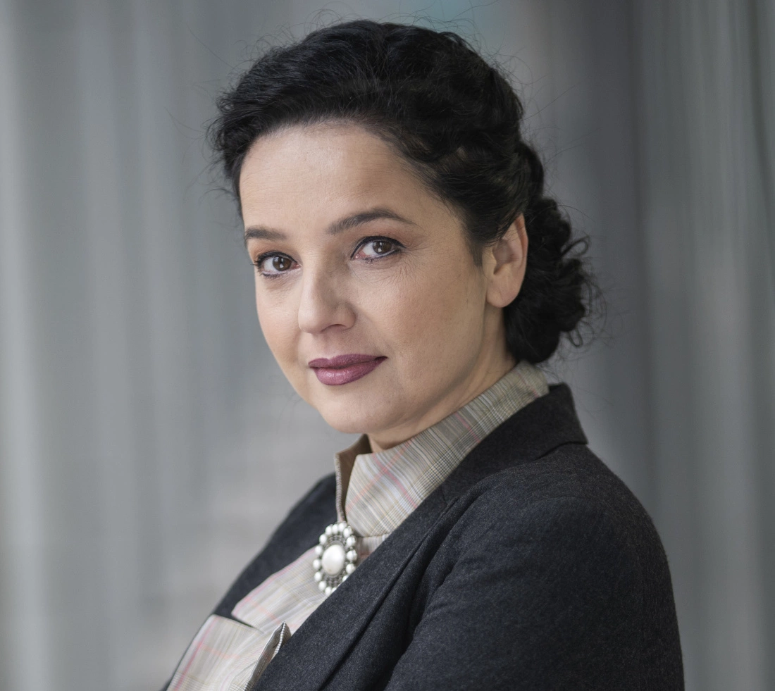 Portret kobiety o ciemnych włosach, w eleganckim stroju, z subtelnym uśmiechem.