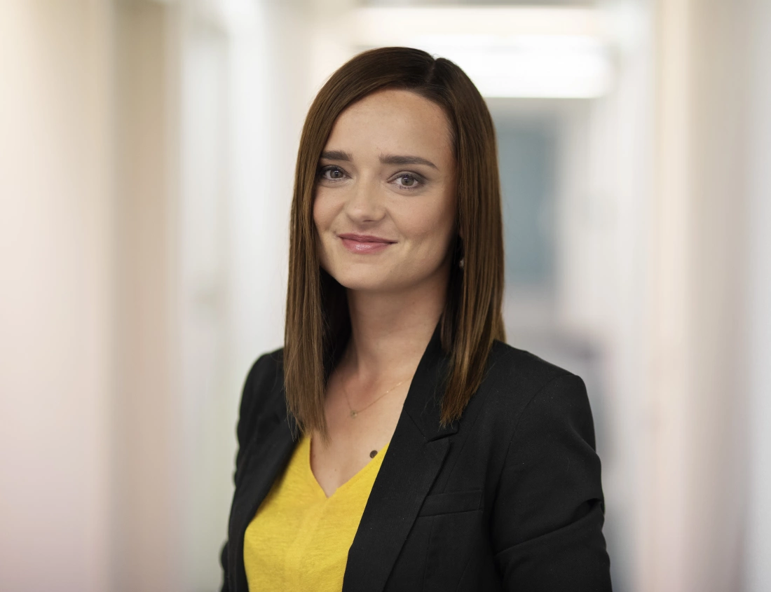 Kobieta w eleganckim stroju, uśmiechająca się na tle biurowego korytarza.