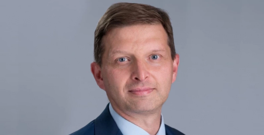 Mężczyzna w granatowym garniturze z jasną koszulą, na tle szarej ściany.