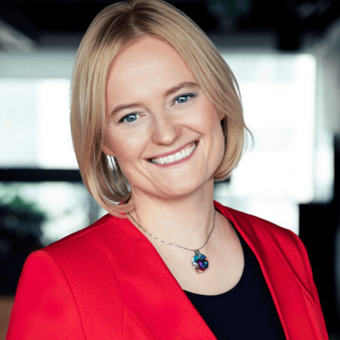 Kobieta z uśmiechem, w czerwonym żakiecie, w nowoczesnym biurze.