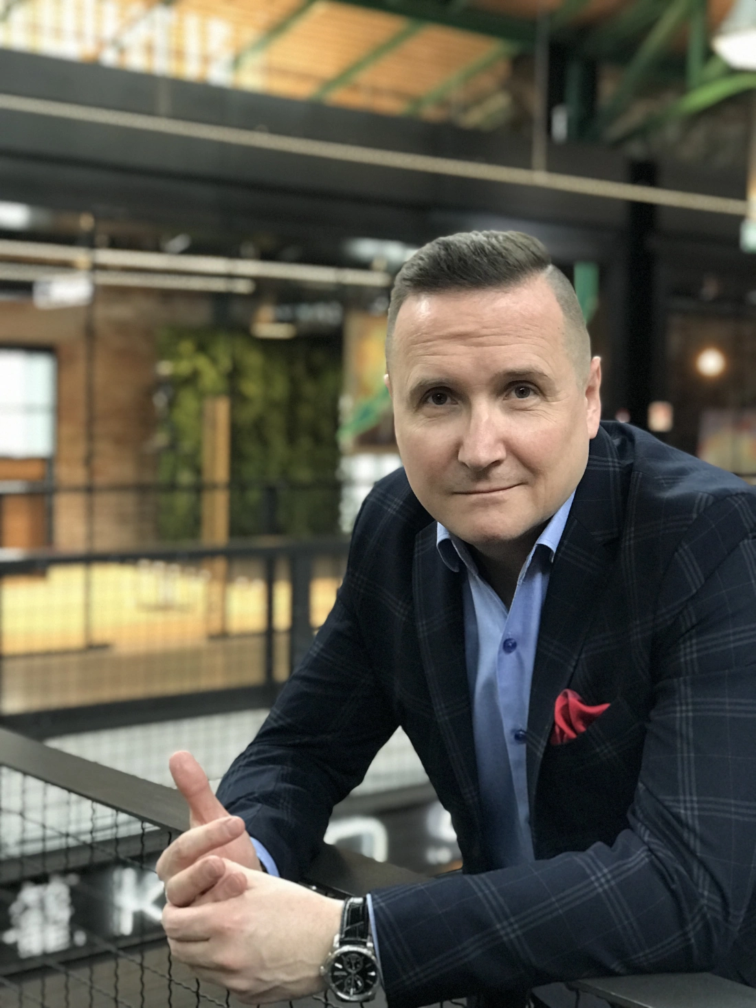 Mężczyzna w eleganckim ubraniu, oparty na barierce, w nowoczesnym wnętrzu.