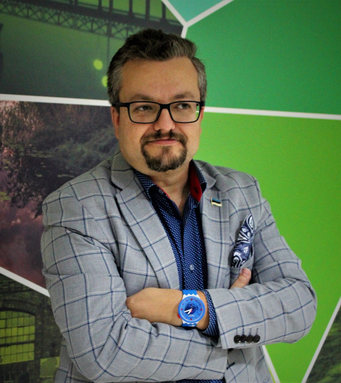 Mężczyzna w szarym garniturze z kratą, z założonymi rękami, uśmiechający się lekko na tle kolorowej ściany.