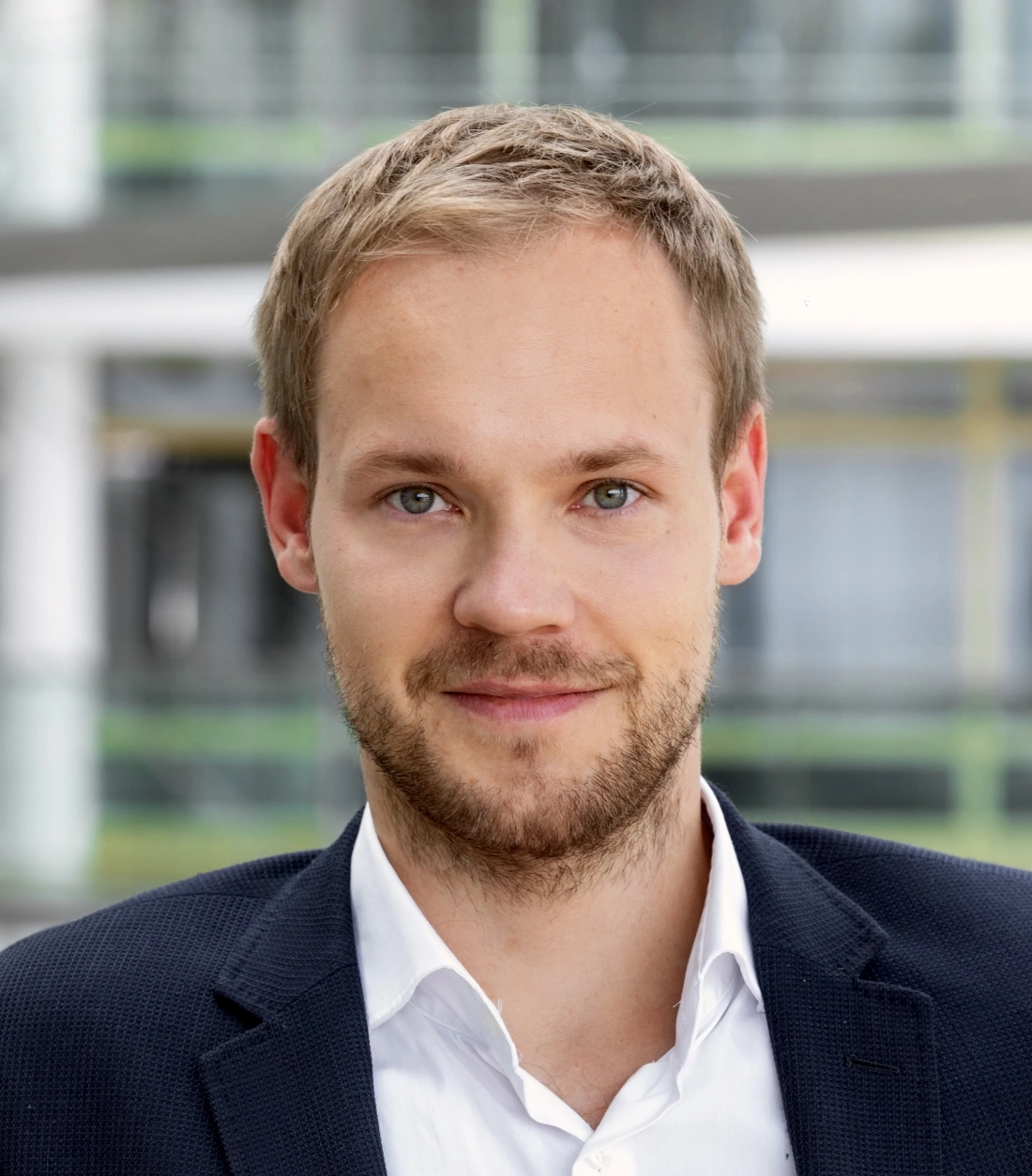 Mężczyzna w garniturze, z krótkimi włosami i brodą, patrzy w stronę obiektywu.