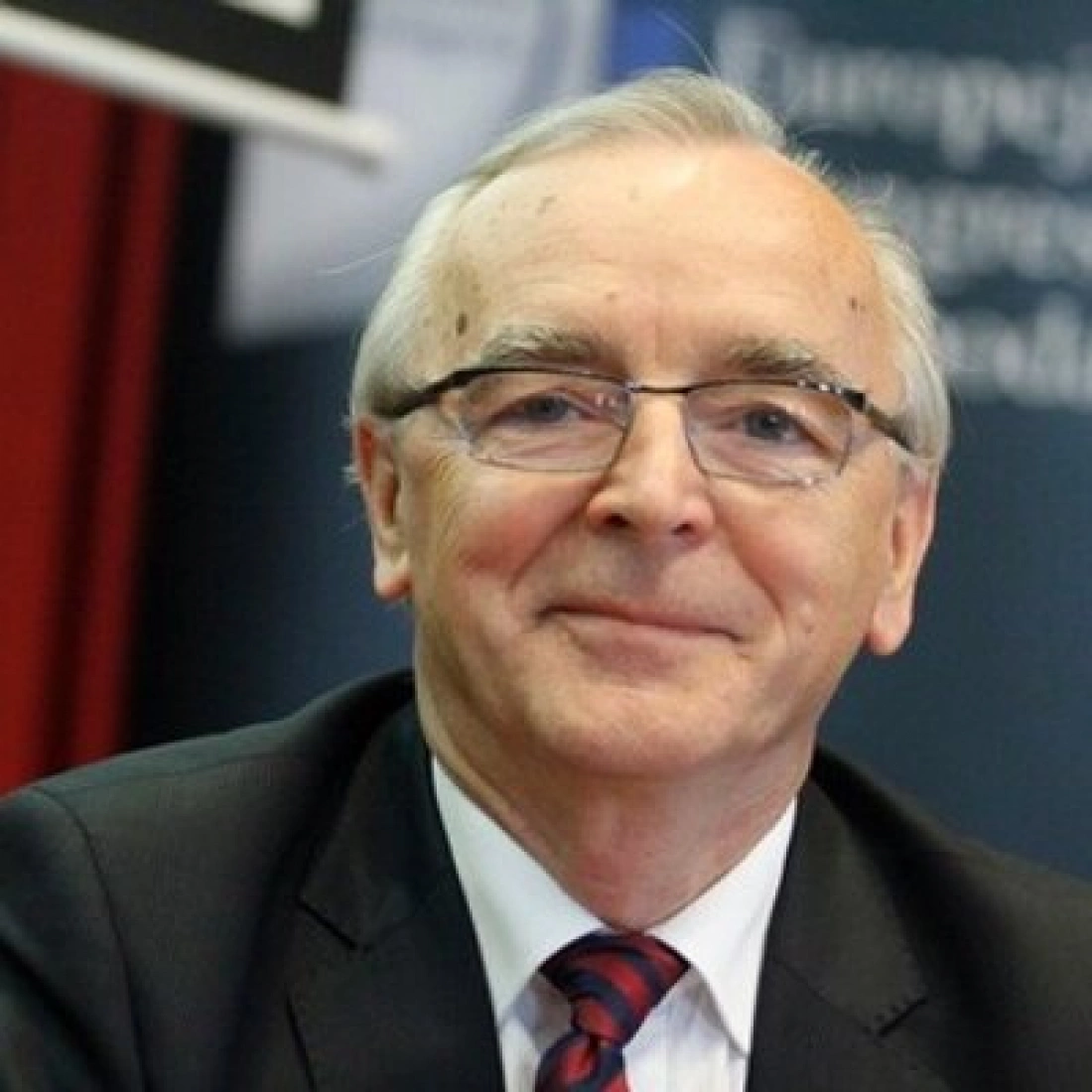 Mężczyzna w okularach, uśmiechający się na tle zasłony.