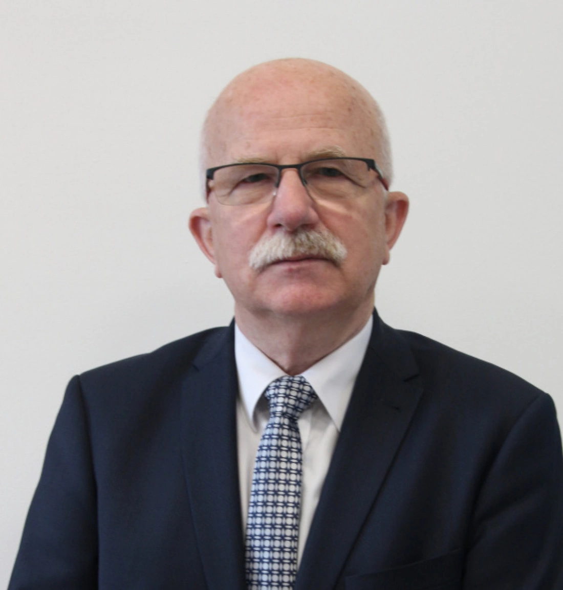 Mężczyzna w garniturze z okularami, stojący na tle białej ściany.