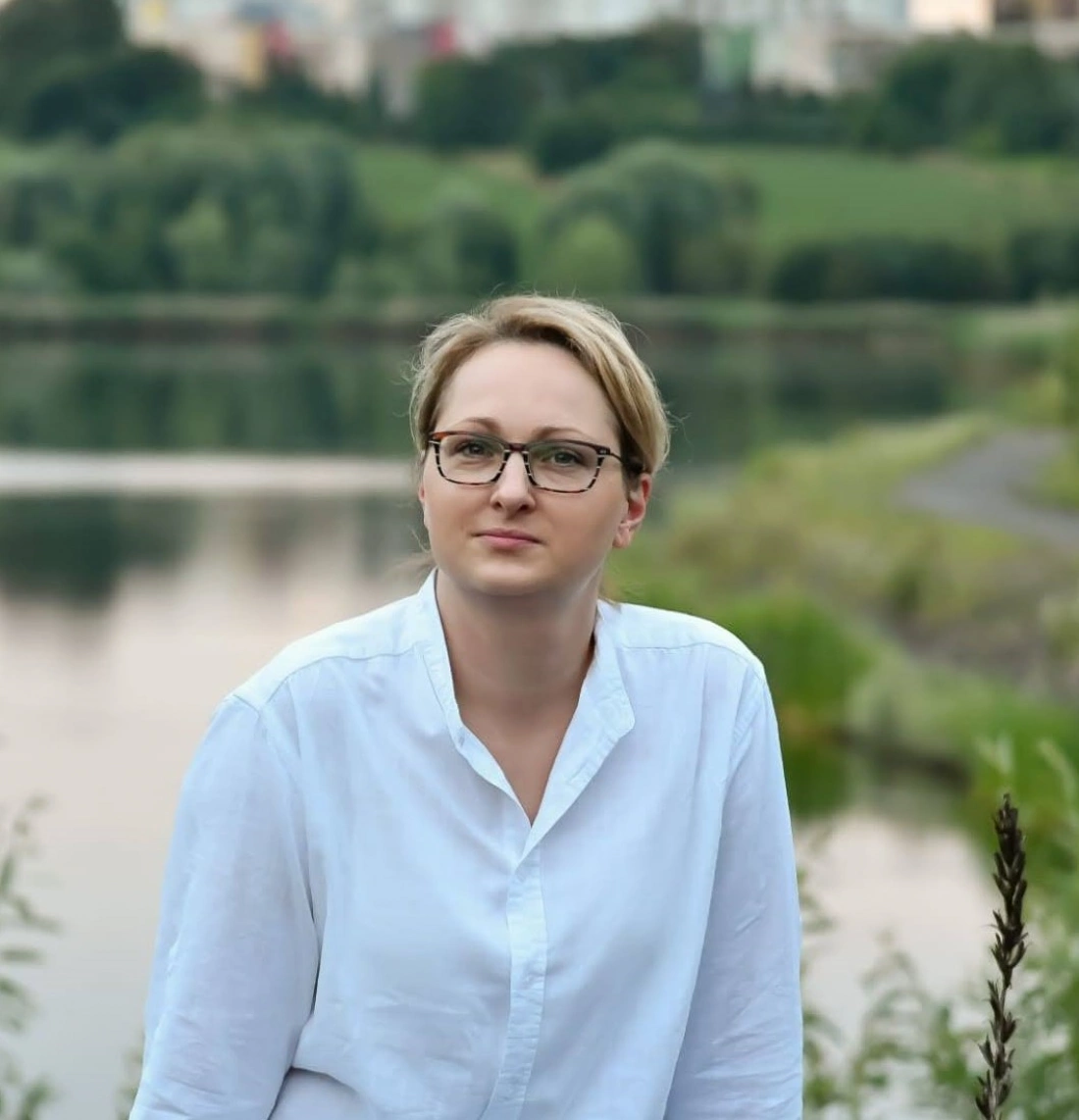 Kobieta w białej koszuli siedzi na brzegu wody, w tle zielone wzgórza.