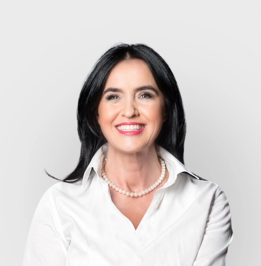 Uśmiechnięta kobieta w białej koszuli i perłowej naszyjniku, stojąca na jasnym tle.