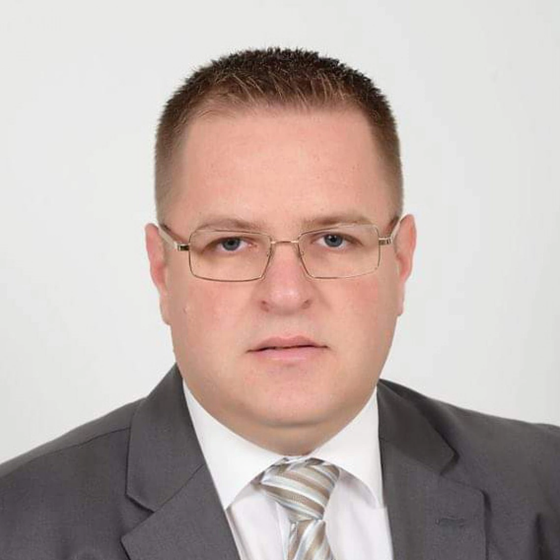 Mężczyzna w garniturze z okularami, poważnym wyrazem twarzy na neutralnym tle.