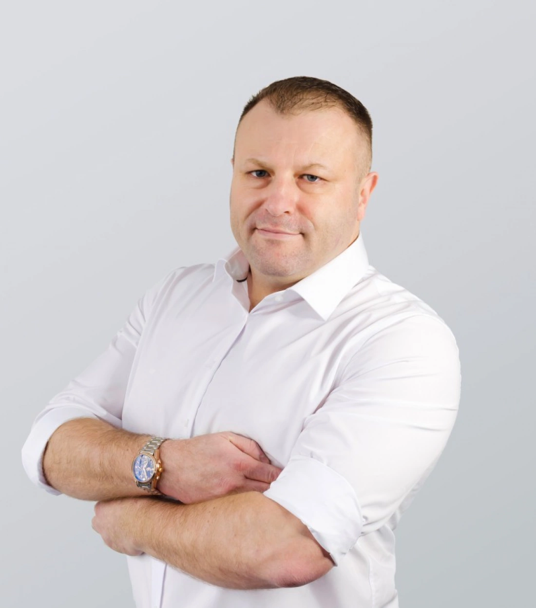 Mężczyzna w białej koszuli z zegarkiem na ręku, pozujący z założonymi rękami.