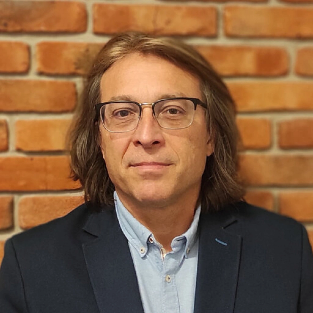 Mężczyzna z długimi włosami, w okularach i eleganckim garniturze, stoi przed ceglanym tłem.