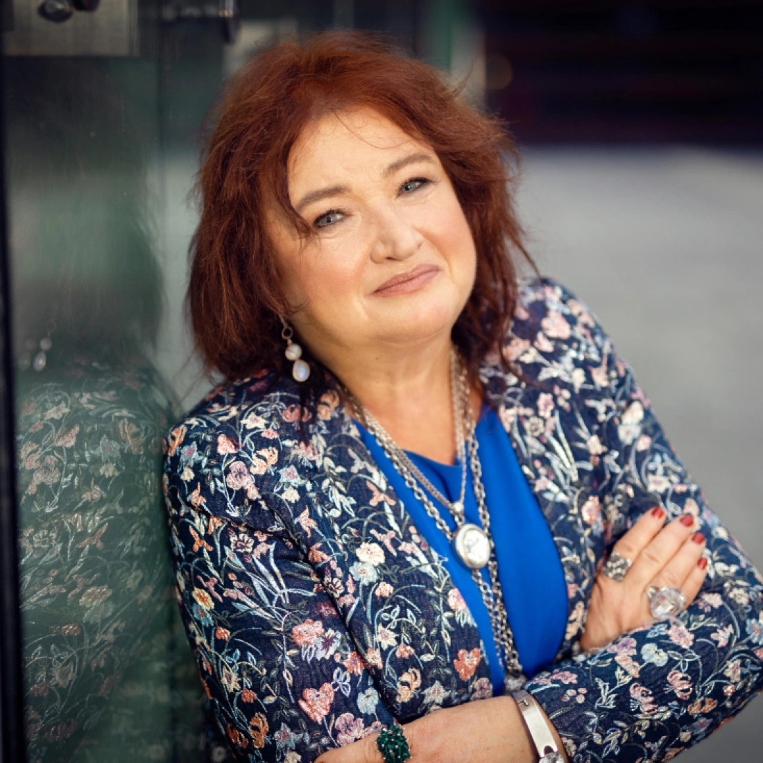 Kobieta o rudych włosach uśmiecha się, opierając się o szklaną ścianę. Ubrana w kwiecisty żakiet i niebieską bluzkę.