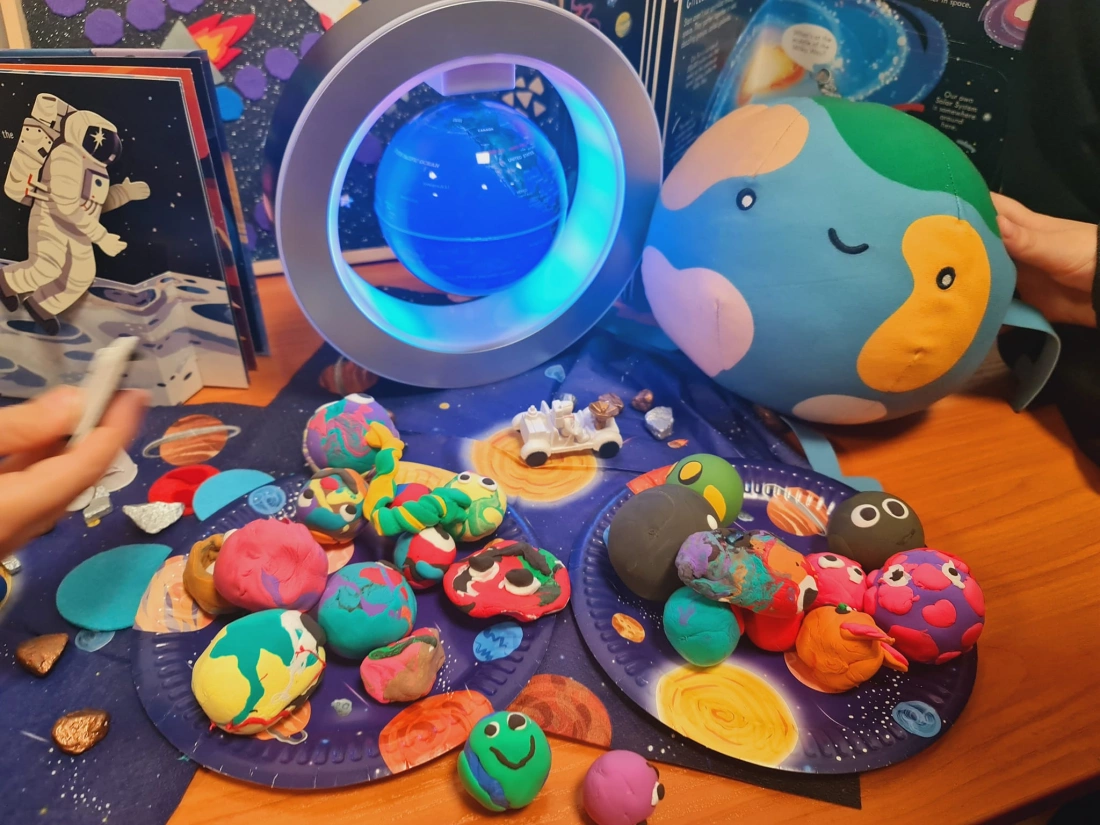 Kolorowe modeliny w kształcie planet leżą na stole obok zabawkowej Ziemi i globusa, z książkami o kosmosie w tle.