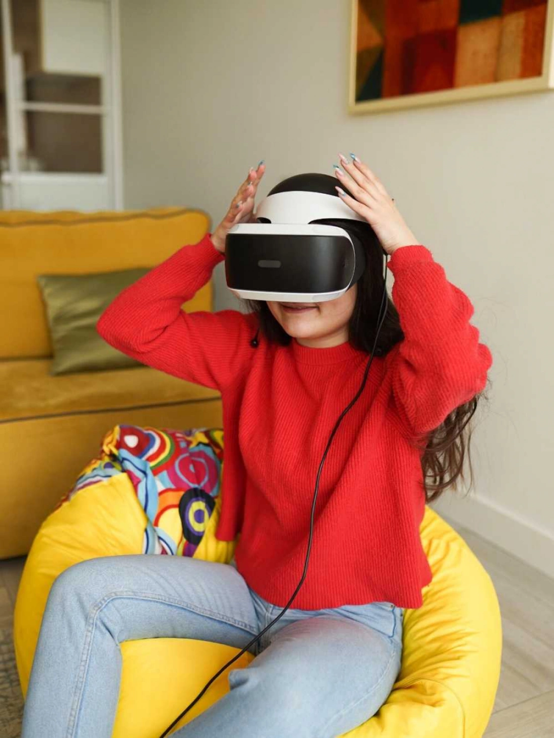 Młoda osoba w czerwonej bluzie nosi gogle VR, siedząc na kolorowym fotelu.