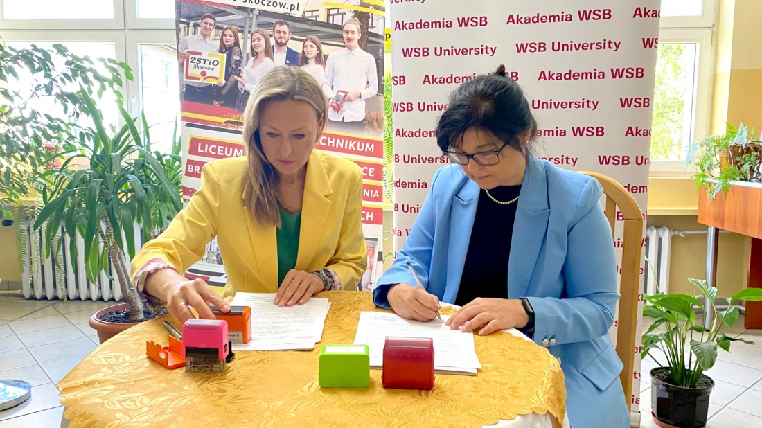 Dwie kobiety siedzą przy stole, podpisując dokumenty, z różnymi pieczątkami na blacie. W tle widoczne plakaty z logo uczelni.