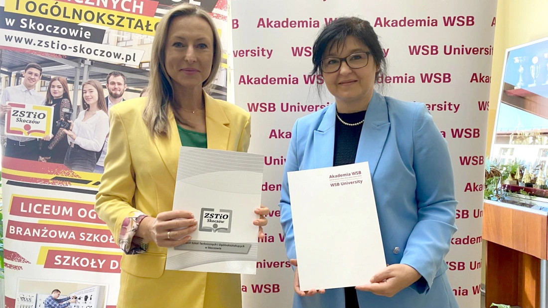 Dwie kobiety trzymają dokumenty w pomieszczeniu z banerem Akademii WSB.