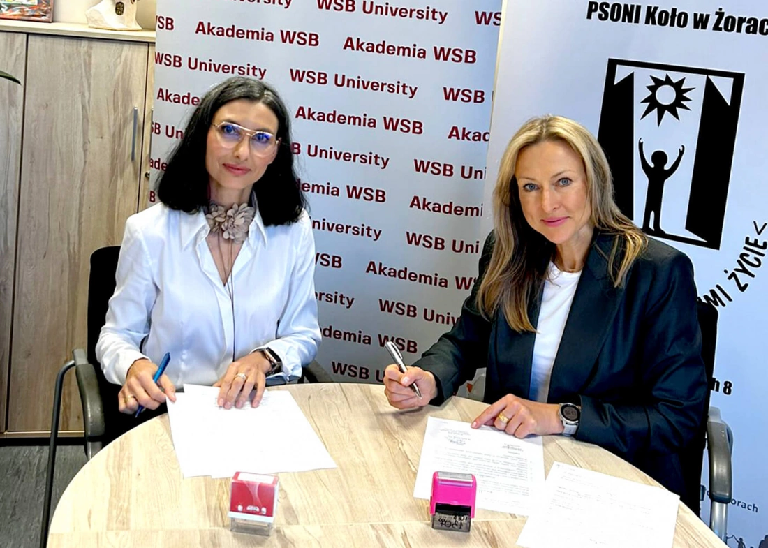 Dwie kobiety siedzą przy stole, podpisując dokumenty, w tle logotypy Akademii WSB.